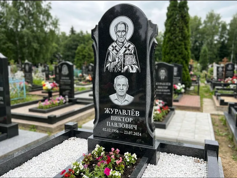 Памятник с гравировкой иконы: стела с образом в полный рост на обороте — гравировка образа Матроны Московской, низкие кусты спиреи в цвету, работа «Лебедева» в Рязани